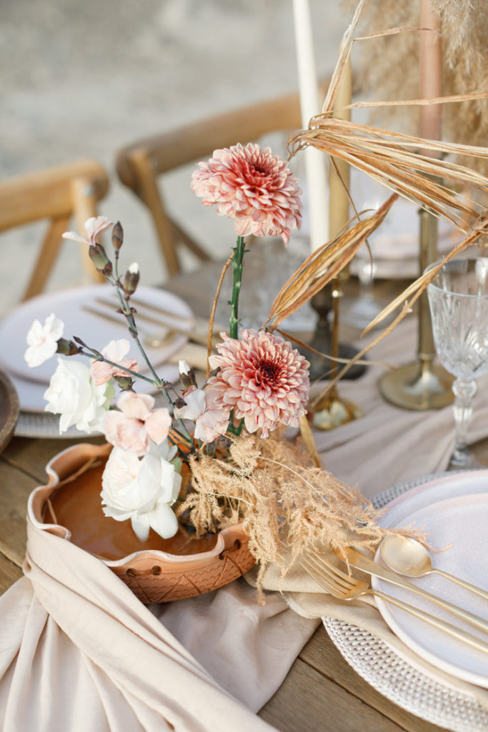 Bohemian Chic wedding style inspiration, close-up of a wedding table scape, minimal floral centerpiece with garden roses, sweet peas, and pampas grass in terracotta pots, brown and brass vintage candleholders, long taper candles, golden cutlery, blush pink plates and white rattan charger plate.