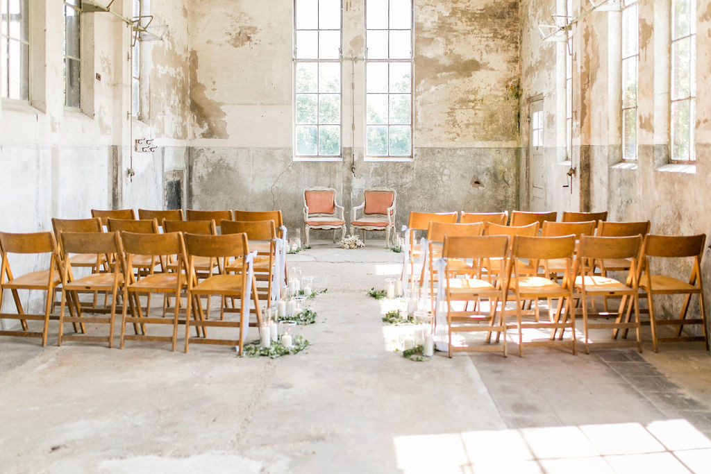 Urban industrial wedding style inspiration, wedding aisle in an old milk factory, high windows, peeling paint, concrete floor, minimal wedding aisle decoration with blue ribbons for the chairs, foliage and pillar candles in glass tubes