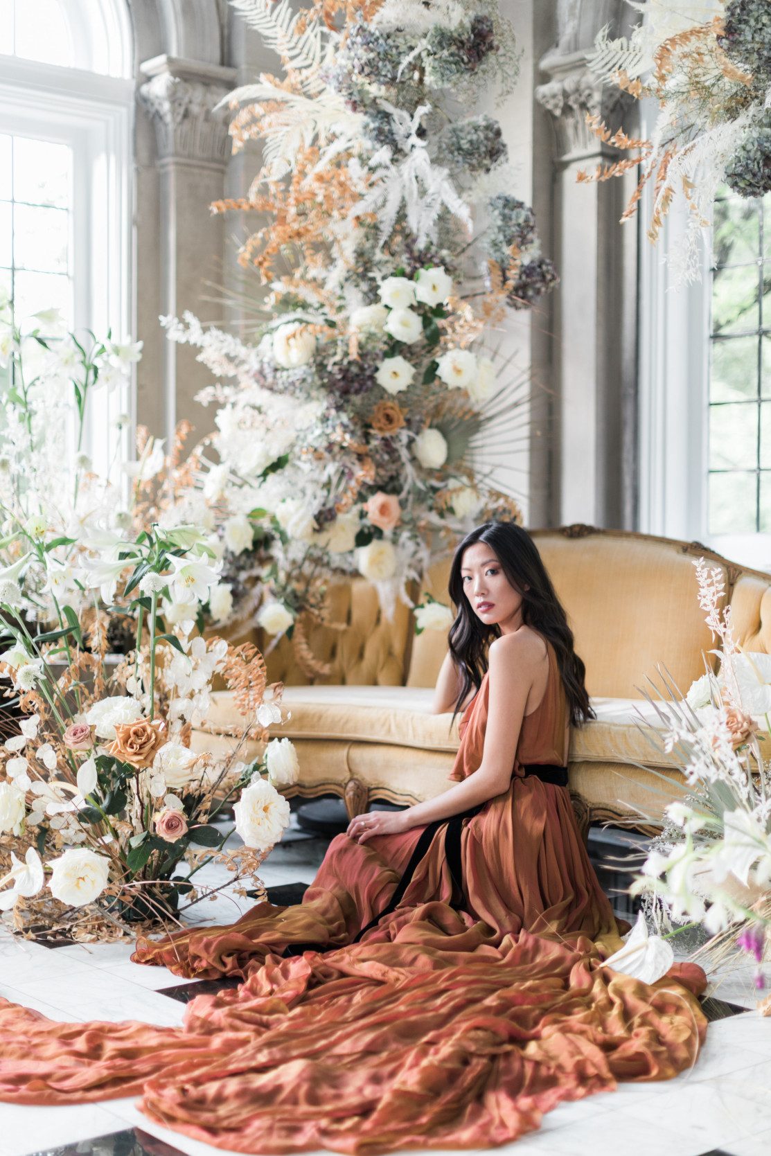 Bride in a rust colored wedding dress sitting in front of a delicate yet decadent floral installation created by Isibeal Studio. Created for a beautiful micro wedding in a historic venue.