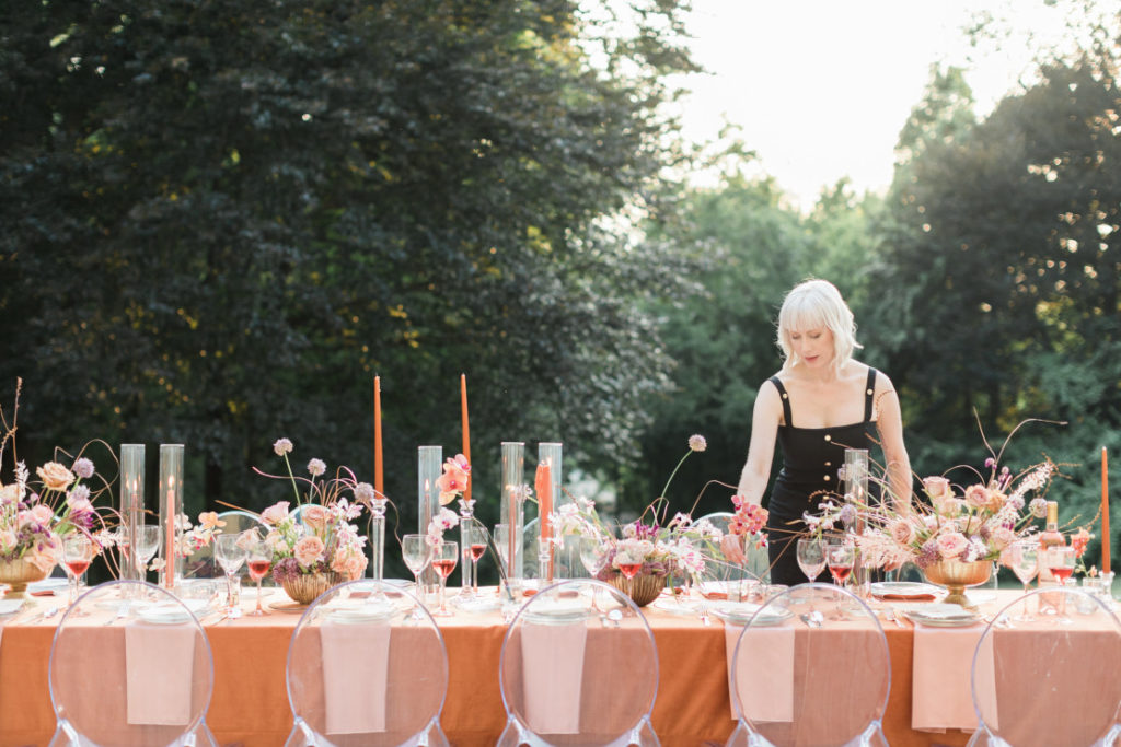 Autumn Keller standing behind an elegant fall al fresco table in orange hues created by Isibeal Studio. Created for a beautiful micro wedding.