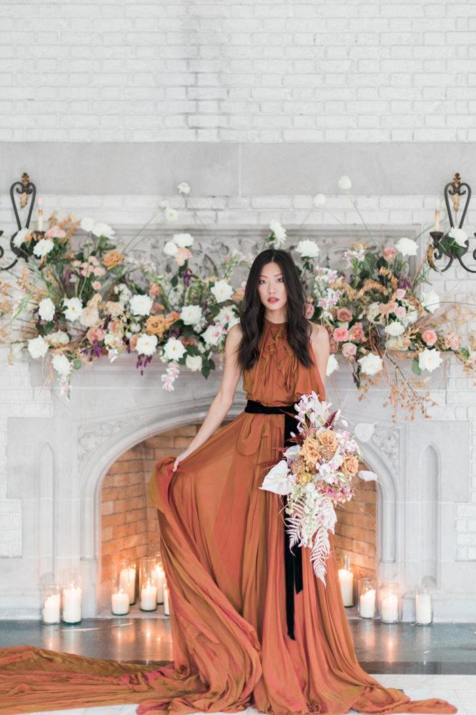 Bride in a rust colored wedding dress in front of a white mantel decorated with lush florals created by Isibeal Studio. Created for a beautiful micro wedding in a historic venue. Decadent yet delicate style.