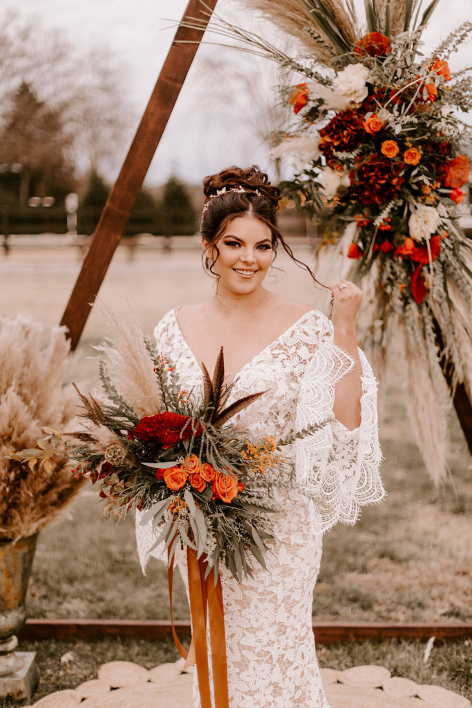 Boho bride standing in front of a triangle wedding arch holding a lush wedding bouquet. The Stylist Abroad, a destination wedding hairstylist, created a gorgeous updo.  
