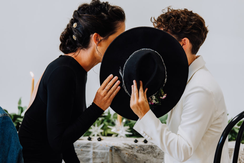 A wedding couple sitting in front of their sweetheart table, covering their faces behind a big black hat. Hairstyles created by The Stylist Abroad, a destination wedding hairstylist. Image by Photos by hfb.