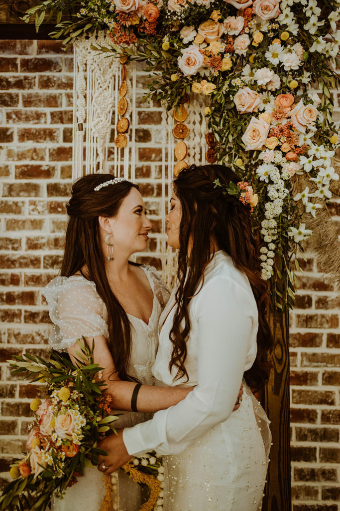 Boho wedding couple looking deeply into each others eyes. Both are dressed in white wedding dresses waering their hair half up half down. Hairstyles created by The Stylist Abroad, a destination wedding hairstylist. Image by The Rose Reflective.