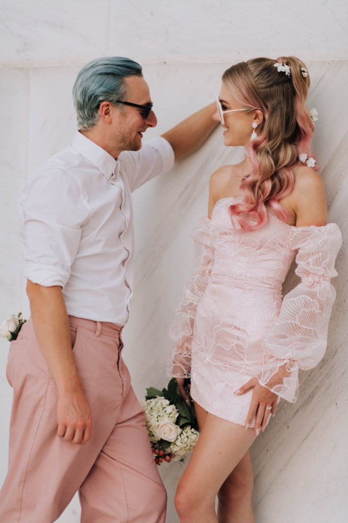 A cool bride and groom. Both dressed in shades of pink, wearing sunglasses. The Stylist Abroad created a creative and fun half up half down hairstyle with cool waves and pink strands. 