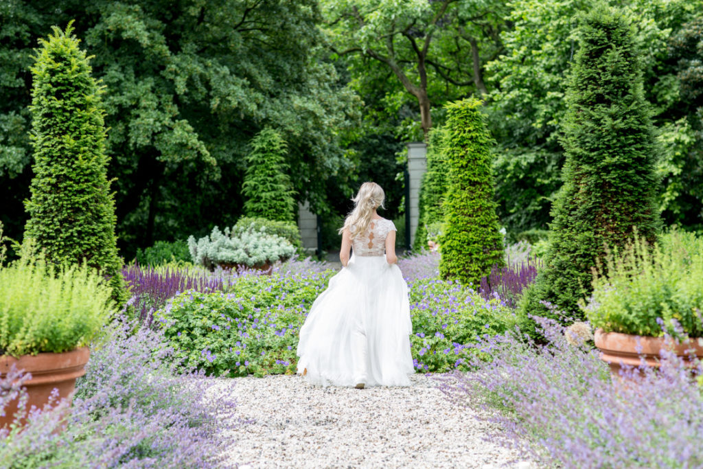 Destination Wedding in Amsterdam:
a bride in the enchanted garden of Huize Frankendael. Violet flowers and lush greenery throughout the garden. Bride wears a gorgeous feminine and romantic wedding dress with a lace back and a flowy, tulle skirt. 
Image by Marielle Kokke Fotografie
#lovelyandplanned

